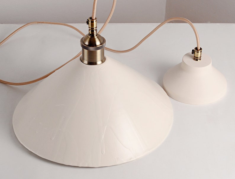 May include: Two white ceramic pendant light fixtures with brass accents and brown cords. The larger fixture has a wide, cone-shaped shade. The smaller fixture has a smaller, round shade.