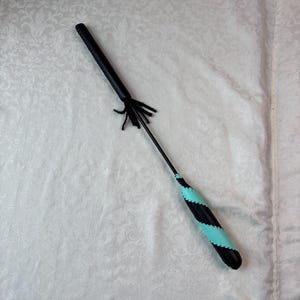 Könnte beinhalten: Eine Reitpeitsche in Schwarz und Türkis mit einem schwarzen Griff und einem türkis-schwarz gestreiften Griff. Die Peitsche hat am Ende des Schafts eine Reihe von schwarzen, lederartigen Strängen.