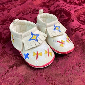 May include: A pair of cream-colored baby moccasins with hand-painted designs. Each shoe features a fringed tongue, a red sole, and painted blue and yellow star and cross motifs. The shoes are designed for infants.