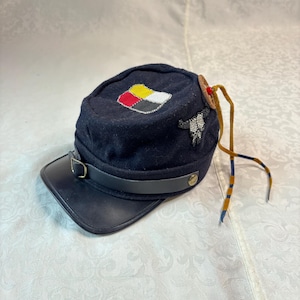 May include: A dark navy blue wool forage cap featuring a black leather visor and a chin strap with a brass buckle. The top is adorned with a beaded patch in red, yellow, white, and black, a beaded bull skull emblem, and a wooden button with leather ties.