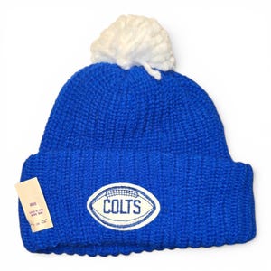 May include: A royal blue knit beanie with a white pom-pom on top and a ribbed cuff. An oval patch on the front features a white football graphic with the text COLTS in blue lettering. A small rectangular paper tag is attached to the side of the cuff.