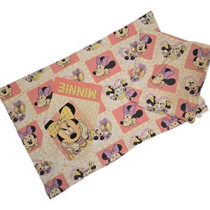 May include: A light pink and white patterned fabric featuring Minnie Mouse. The design includes images of Minnie Mouse in various poses, set against pink squares and polka dots. The word "MINNIE" is also visible.