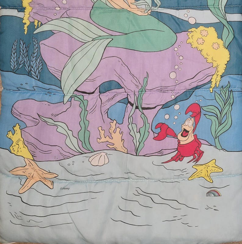Może przedstawiać: Kolorowa ilustracja przedstawiająca syrenę z zielonymi włosami i ogonem, siedzącą na fioletowej skale. Przedstawiono r&oacute;wnież czerwonego kraba, ż&oacute;łte rozgwiazdy i r&oacute;żne rośliny morskie. Tło jest jasnoniebieskie.
