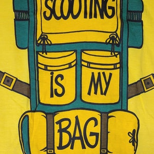 May include: A yellow t-shirt with a green and brown backpack design. The backpack has two pockets with the words "is" and "my" on them. The bottom of the backpack has the word "BAG" on it. The text on the shirt reads "Scooting is my bag."