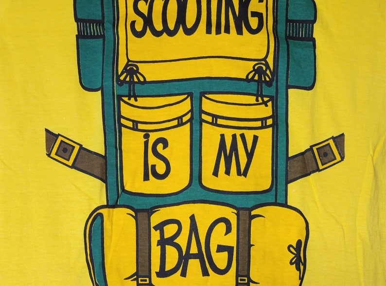 May include: A yellow t-shirt with a green and brown backpack design. The backpack has two pockets with the words "is" and "my" on them. The bottom of the backpack has the word "BAG" on it. The text on the shirt reads "Scooting is my bag."