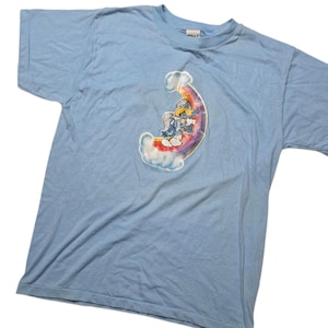 May include: Light blue t-shirt featuring a colorful graphic of Smurfs riding a rainbow. The graphic includes clouds and a rainbow arch. The shirt is made of a soft material and has short sleeves.