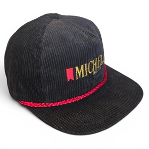May include: A black corduroy baseball cap featuring a flat brim and a red braided rope trim across the base of the crown. The front displays the embroidered text MICHELOB in gold lettering, accompanied by a small red vertical banner icon to the left.