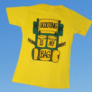 May include: Yellow t-shirt with a green and brown cartoon backpack on the back. The backpack has the text "SCOUTING IS MY BAG" on it.