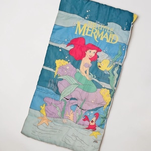 Może przedstawiać: Niebieski śpiw&oacute;r z kolorową ilustracją Arielki z Małej Syrenki Disneya. Projekt zawiera Ariel, Florka, Sebastiana i elementy podwodne. Śpiw&oacute;r jest prostokątny i ma pikowaną fakturę.