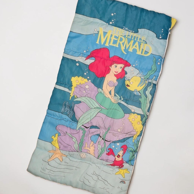 Może przedstawiać: Niebieski śpiw&oacute;r z kolorową ilustracją Arielki z Małej Syrenki Disneya. Projekt zawiera Ariel, Florka, Sebastiana i elementy podwodne. Śpiw&oacute;r jest prostokątny i ma pikowaną fakturę.