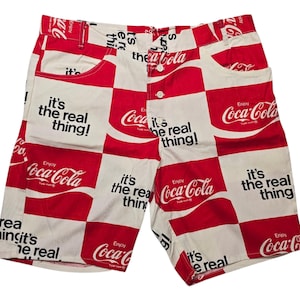 May include: White and red checkered shorts featuring the Coca-Cola logo and the phrase "it's the real thing!". The shorts have two front pockets and a button closure.