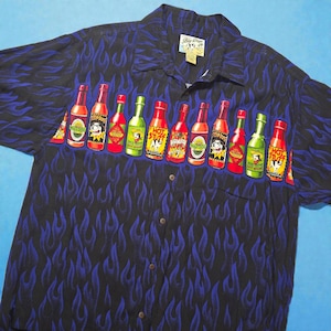 May include: Black button-down shirt with a blue flame pattern. The shirt features a row of colorful hot sauce bottles with cartoon dog characters. The shirt has a collared neckline and a chest pocket.