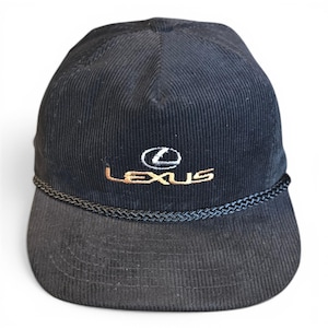 May include: A black corduroy baseball cap featuring the Lexus logo embroidered in gold and white thread on the front panel. The cap includes a decorative braided black cord trim positioned horizontally across the base of the crown above the brim.
