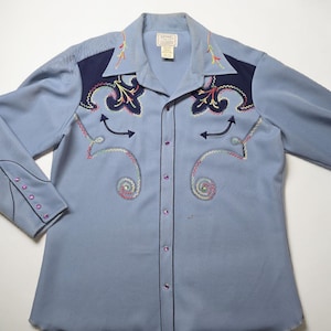 May include: A light blue long-sleeve shirt with dark blue shoulder panels. The shirt features colorful embroidered designs, including swirls, arrows, and floral patterns. It has a button-down front and cuffs with purple buttons.