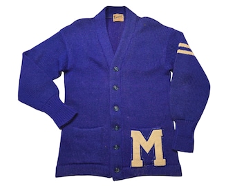 May include: A vintage purple cardigan sweater with a large felt letter "M" on the front pocket. The sweater has two white stripes on each sleeve and a button-up front.