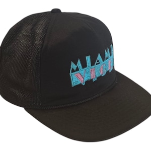 May include: Black trucker hat with a mesh back and a curved brim. The front of the hat features the embroidered text "MIAMI VICE" in teal and pink. The hat has a button on top.