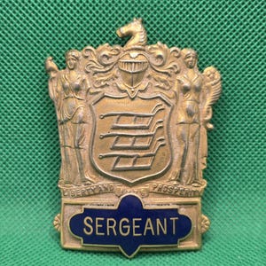 May include: A vintage brass Sergeant badge with a blue enamel banner. The badge features a crest with a horse, two female figures, and the words "Liberty and Prosperity". The badge is likely from a military or law enforcement organization.