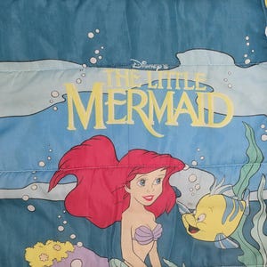 Może przedstawiać: Niebiesko-biały materiał z logo Małej Syrenki Disneya i ilustracjami Arielki i Florka. Arielka ma rude włosy i fioletowy biustonosz z muszelek. Florek jest ż&oacute;łty z niebieskimi paskami. Tło przedstawia podwodną scenę.