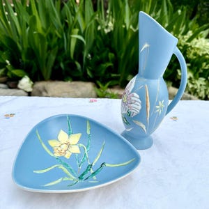 Peut inclure: Un pichet en céramique bleu clair et un plat triangulaire assorti. Le pichet présente un motif floral peint avec une anse et un bord incliné. Le plat affiche une jonquille jaune unique avec des tiges et des feuilles vertes peintes.