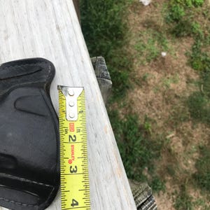 May include: A black leather holster with a stitched design. A yellow Stanley measuring tape is next to the holster, showing a measurement of approximately 7.6 cm.