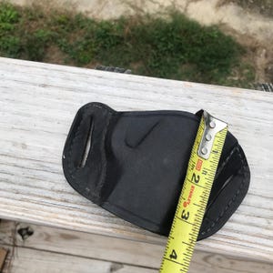 May include: A black leather holster with a curved shape, stitched edges, and cutouts. A yellow measuring tape shows the holster is approximately 10 cm long. The holster is on a wooden surface.