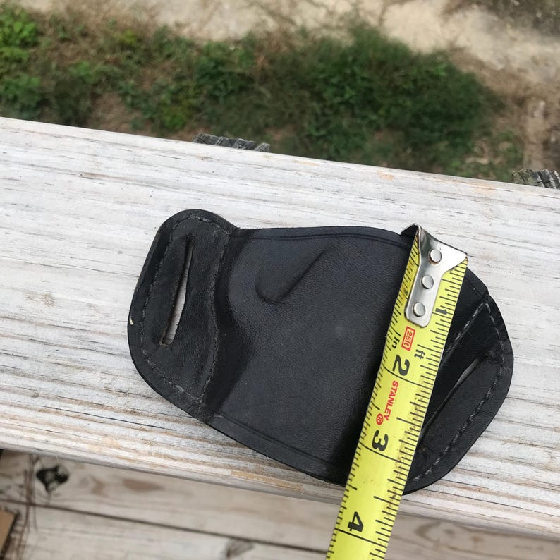 May include: A black leather holster with a curved shape, stitched edges, and cutouts. A yellow measuring tape shows the holster is approximately 10 cm long. The holster is on a wooden surface.