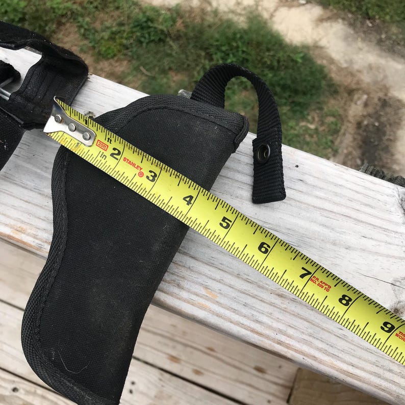 May include: A black nylon holster with a loop handle and snap closure, measured with a yellow Stanley tape measure. The holster is approximately 13 cm long.