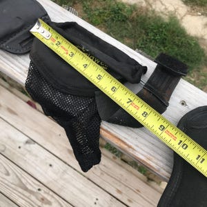 May include: Black equestrian equipment, including a mesh-lined boot protector, a leather-like strap, and a black pad. A yellow measuring tape shows the boot protector is approximately 18 cm long.