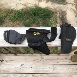 May include: Three black gun holsters are arranged on a wooden surface. One is leather, the others are nylon. One holster has the text "Caldwell Shooting Supplies" printed on it. The holsters are for carrying firearms.