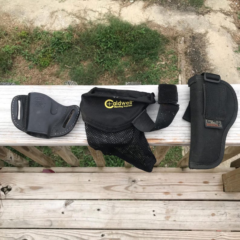 May include: Three black gun holsters are arranged on a wooden surface. One is leather, the others are nylon. One holster has the text "Caldwell Shooting Supplies" printed on it. The holsters are for carrying firearms.