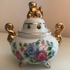 May include: A white porcelain incense burner with a gold trim and floral design. The lid features a gold figurine of a sitting animal.