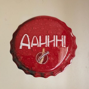 May include: A red bottle cap with the word "AAHHH!" in white block letters. The cap has a silver and gold graphic in the center and is covered in water droplets. The edges of the cap are crimped.