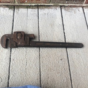 May include: A rusty, vintage pipe wrench with a long handle and adjustable jaws. The wrench is dark brown and shows signs of wear. The text "FREMONT N.Y. U.S.A." is visible on the handle. The tool is designed for gripping and turning pipes.