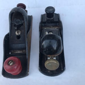 May include: Two vintage hand planes. One has a red knob and the other has a black knob. Both are black with metal blades and are used for woodworking. The planes are set against a white background.