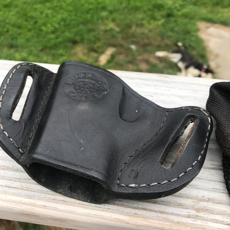 May include: Black leather holster with white stitching. The holster features two belt slots and a stamped logo reading "Bulldog Cases". Designed to securely hold a firearm.