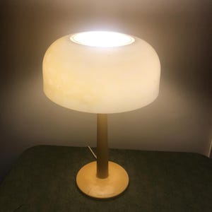 May include: A yellow table lamp with a white, round shade. The lamp is turned on and the light is shining through the shade.