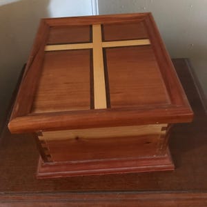 May include: A wooden box with a cross inlay on the lid. The box is made of light and dark wood and has a natural finish.