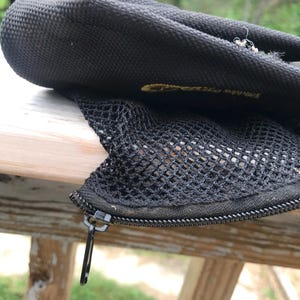 May include: Black mesh and nylon bag with a zip closure. The bag has a textured surface and a small metal zip pull. The bag appears to be partially open, revealing the mesh interior. The bag is resting on a wooden surface.