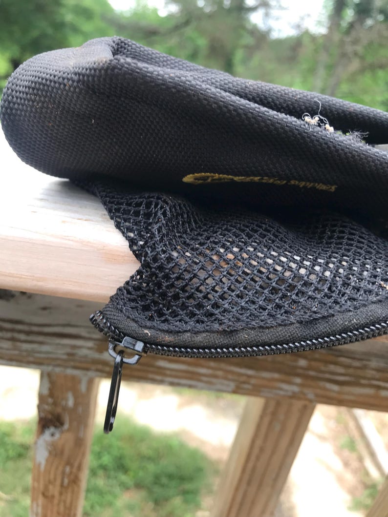 May include: Black mesh and nylon bag with a zip closure. The bag has a textured surface and a small metal zip pull. The bag appears to be partially open, revealing the mesh interior. The bag is resting on a wooden surface.