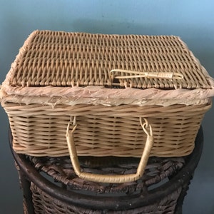 May include: A woven wicker basket with a handle. The basket is light brown and has a natural woven texture. The basket is sitting on a brown wicker stool.