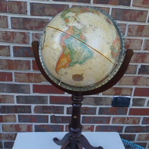 May include: A vintage globe with a cream-colored surface, displaying continents in various colors, mounted on a dark brown wooden stand. The globe is held by a dark wooden frame. The stand has a decorative base.