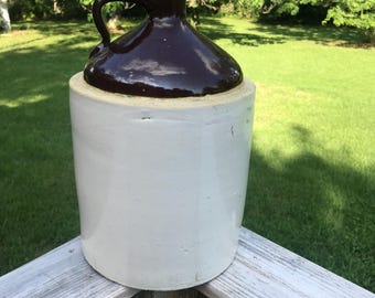 May include: A vintage stoneware jug with a dark brown, glossy top and handle, and a cylindrical, off-white body. The jug is likely used for storing liquids and has a rustic, handcrafted appearance. The jug is sitting on a wooden surface.