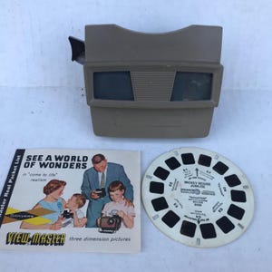 May include: An antique View-Master viewer in a tan color, with a packet list and a Mickey Mouse Jubilee reel. The packet list features a family looking at a View-Master. The reel is white with black frames.