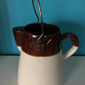 May include: A white and brown ceramic pitcher with a metal handle. The pitcher has a wide mouth and a rounded body.