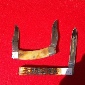 May include: Three pocket knives with open blades are displayed against a red background. The knives have bone-colored handles and silver blades. One blade reads "Bob Younger", and another reads "LITTLE PILLBUSTER".