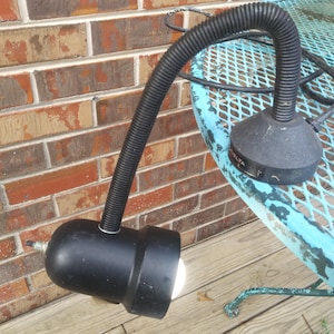 May include: A black Delta gooseneck lamp with a flexible, ribbed neck. The lamp is positioned on a light blue metal table and has a bright light emanating from the bulb. The lamp is on a wooden deck with a brick wall in the background.