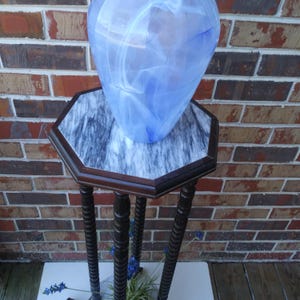 May include: A large, blue and white swirled glass vase sits on a marble-topped, dark wood hexagonal table. The table has three turned legs. The background is a brick wall.