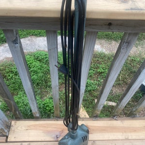 May include: A gray electric water pump with a black power cord and plug. The pump is hanging from a metal hook. The pump is sitting on a wooden deck.