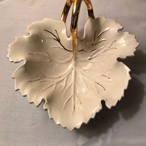 May include: White ceramic leaf-shaped serving dish with a gold handle. The dish has a gold leaf design.