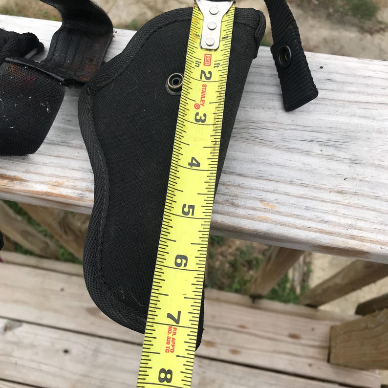 May include: Black nylon holster, approximately 20 cm long, with a black strap and metal rivets. A yellow tape measure shows the holster's length. The holster is designed to hold a firearm.